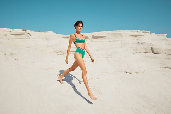 Woman in a green kiini bikini walking on a sandy beach with white cliffs in the background